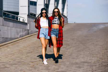 Two young happy hipster girls in sunglasses having fun, smiling, laughing, jumping, walking outdoor on the street, summer relax conceptの写真素材