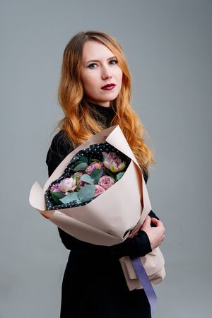 Portrait of red-haired young girl florist with bouquet of flowers in dusty pink package in the hands on light background. Rose, hyacinth, anemone, eucalyptus.の写真素材