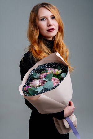 Portrait of red-haired young girl florist with bouquet of flowers in dusty pink package in the hands on light background. Rose, hyacinth, anemone, eucalyptus.の写真素材