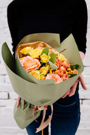 Bouquet of flowers in pistachio-colored package in the hands of a woman florist on background a white brick wall. Rosa, stahis, ilexの写真素材