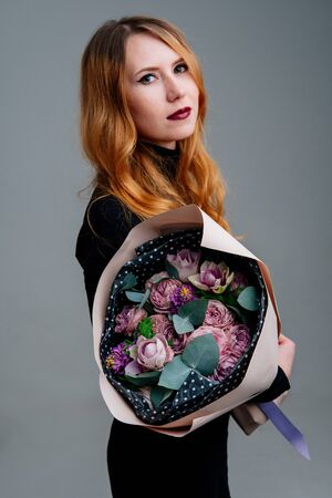 Portrait of red-haired young girl florist with bouquet of flowers in dusty pink package in the hands on light background. Rose, hyacinth, anemone, eucalyptus.の写真素材