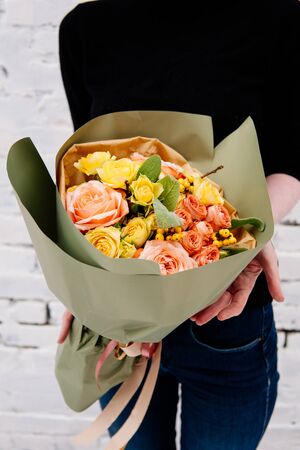 Bouquet of flowers in pistachio-colored package in the hands of a woman florist on background a white brick wall. Rosa, stahis, ilexの写真素材