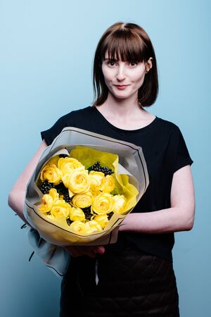 Portrait of young female florist with bouquet of flowers in transparent package in the hands on a blue background . Rosa spray Catalina, rhamnusの写真素材