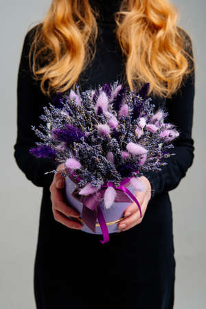 Bouquet in lilac hat box of dried flowers in the hands of red-haired girl florist on light...