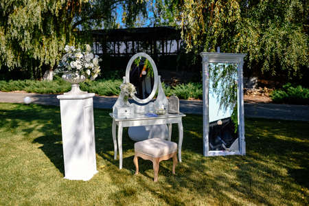 Place for a photo for the bride. White luxury chair stands before a mirror and wedding flower arrangements on the green lawn outsideの写真素材