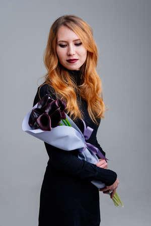 Portrait of red-haired young girl florist with bouquet of calla lilies in lilac foamiran package in the hands on light background.の写真素材