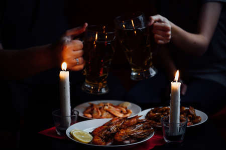 two hands, two clink glasses with beer, two candles, shrimps and lobster with lemon on a red tableclothの写真素材