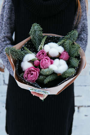Bouquet of flowers and fir branches and cottonwood boxes in peach package in the hands of woman florist in knitted sweater on background white brick wall.の写真素材