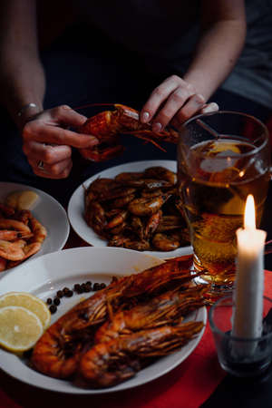 Woman's hand peeled grilled lobster on a red tablecloth, plates with shrimps, a glass of beer, a candleの写真素材