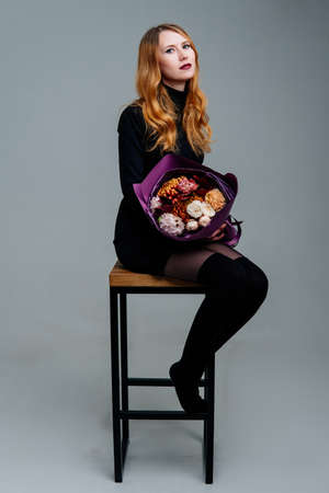 Portrait of red-haired young girl florist sitting on chair with bouquet of flowers in purple package on light background. Rose, chrysanthemum, carnation, cotinus.の写真素材