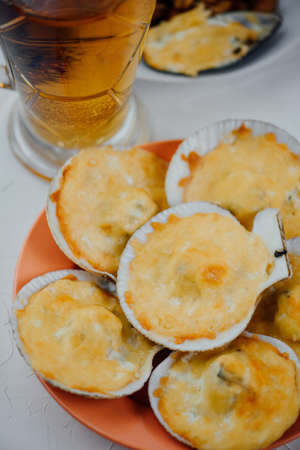 scallops in a shell under the cheese in an orange plate and a glass of beer on a light background