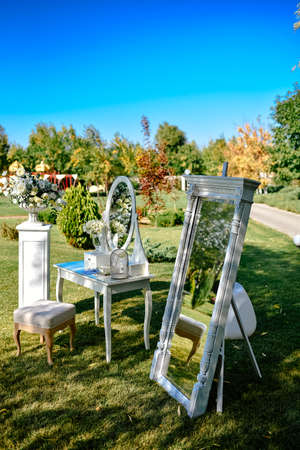 Place for a photo for the bride. White luxury chair stands before a mirror and wedding flower arrangements on the green lawn outsideの写真素材