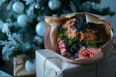 Bouquet of flowers in peach package on gift box wrapped in craft paper on the background of Christmas tree. Rosa, carnation, eryngium, eucalyptus.の写真素材