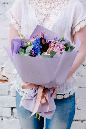 A bouquet of flowers, packed in purple paper in the hands of a woman on a light background. Bouquet of Eucalyptus, Carnation, Hydrangea, Rose, Clematis, Cotinus, Gladiolusの写真素材