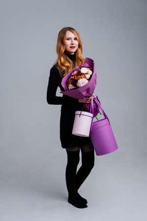 Portrait of red-haired young girl florist bouquet of flowers in purple package and hat gift boxes in the hands on light background. Rose, chrysanthemum, carnation, cotinus.の写真素材