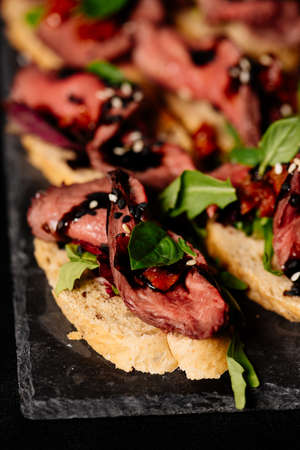 Bruschetta with baked beef, arugula and balsamic sauce on black tableの写真素材