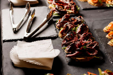 Bruschetta with baked beef, arugula and balsamic sauce on black table with white napkins and kitchen tongsの写真素材