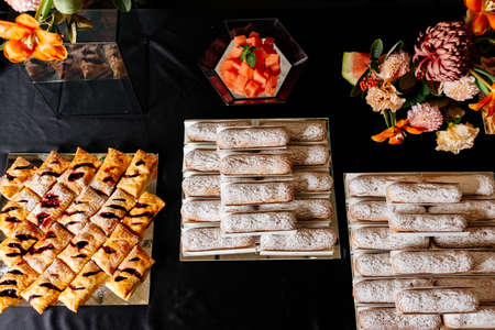 Tasty vanilla eclairs individually wrapped sprinkled with powdered sugar and sweet puff buns with jam sprinkled with powdered sugar on black background and orange flowers bouquet on black tableの写真素材