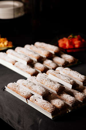 Tasty vanilla eclairs individually wrapped sprinkled with powdered sugar and orange flowers bouquet on black tableの写真素材
