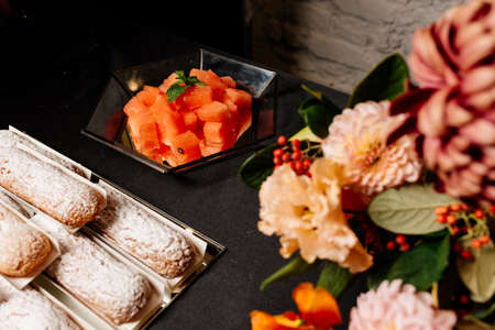 Tasty vanilla eclairs individually wrapped sprinkled with powdered sugar and bouquet of flowers with tulip, dahlia, chrysanthemum, eustoma in orange tones and watermelon on dark backgroundの写真素材