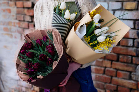 Various bouquets of tulips in beige, burgundy, striped black and pistachio-colored package in the hands of girl florist in knitted pullover on brick wall background.の写真素材
