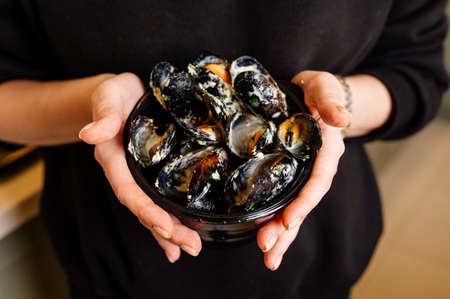Black plate with delicious fried mussels in shells with creamy sauce in hands of woman in black sweaterの写真素材