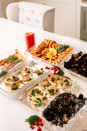 Luxury sea scallop in shell in becon with greens in plastic box and delicious fried langoustines with dill and lemon in plastic box and mussels in shells with cheese and fried shrimps in plastic box and red berries and gold and red burning candles on festive tableの写真素材