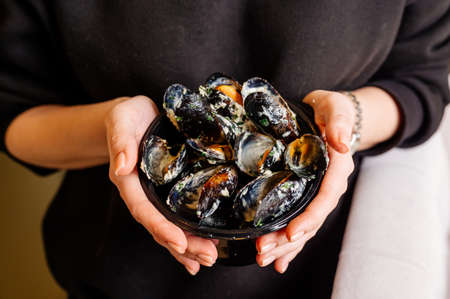 Black plate with delicious fried mussels in shells with creamy sauce in hands of woman in black sweaterの写真素材