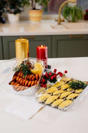 Mussels in shells baked with cheese in a plastic box and fried shrimps in a plastic box and red berries and gold and red burning candles on kitchen tableの写真素材