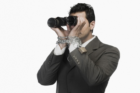 Businessman locked in chains and looking through binocularsの写真素材