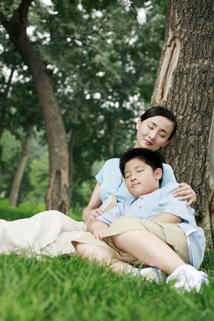 Woman and boy leaning against a tree with their eyes closedの写真素材