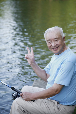 Senior man fishing at the lake, showing peace signの写真素材