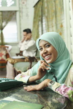 Woman smiling while talking on the phone, man using laptop in the backgroundの写真素材