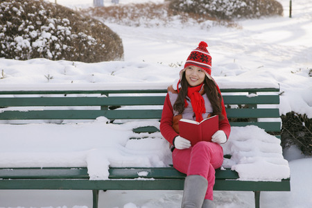 Woman in warm clothing reading book on a snowy bench の写真素材
