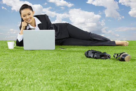 A relaxed young Asian businesswoman lying on grass using her laptopの写真素材