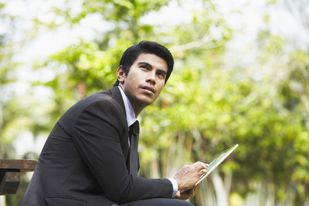 Young Asian businessman sitting on a park bench using a digital tabletの写真素材
