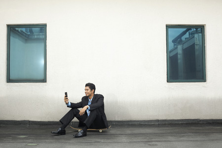 Young businessman sitting on skateboard, text messaging on phoneの写真素材
