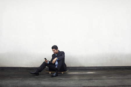 Young businessman sitting on skateboard, text messaging on phoneの写真素材