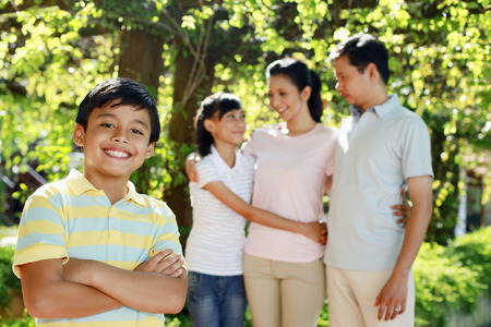 Boy standing with arms crossed, his family in the backgroundの写真素材