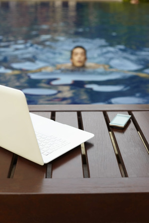 Laptop and mobile phone on lounge chair, man swimming in the poolの写真素材
