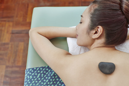A young woman lying on her front relaxing with massage stone on her backの写真素材