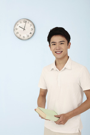 Man smiling while holding book, clock on the wallの写真素材