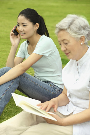 Senior woman reading book while woman is talking on the phoneの写真素材