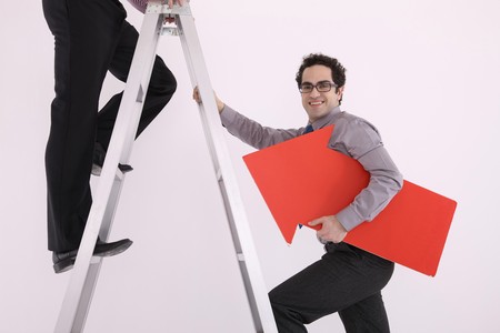 Businessman standing on the ladder, another businessman climbing up while holding arrow signの写真素材