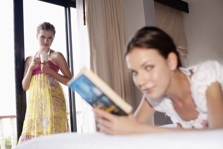 Woman reading book on bed, her friend holding a cup in the backgroundの写真素材
