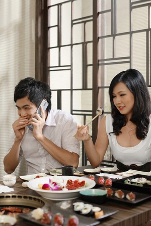 Woman picking up sushi with chopsticks, man giggling while talking on the phoneの写真素材
