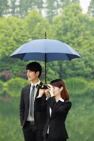 Businesswoman looking through binoculars, businessman shielding her with an umbrellaの写真素材