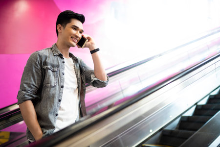 Young Asian man talking on phone while standing on escalator