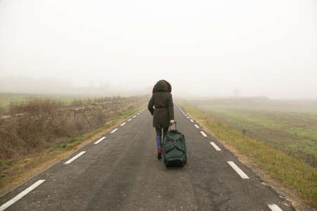 Unrecognizable woman, walking with suitcase in hand on a cold and foggy mountain oh the roadの写真素材