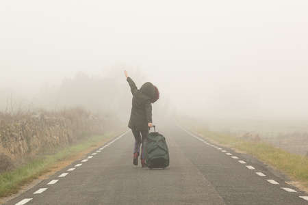 Unrecognizable woman, walking with suitcase in hand on a cold and foggy mountain oh the roadの写真素材